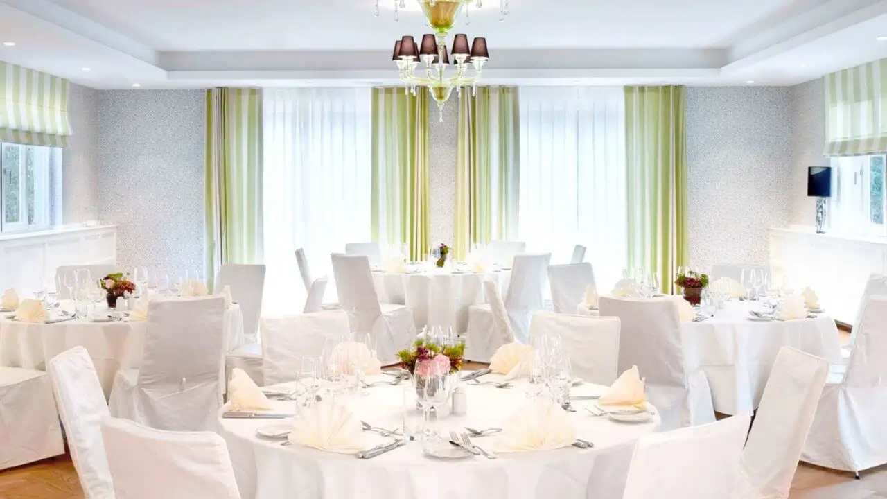 Feines Restaurant mit gedeckten Tischen, weißen Stoffhussen und grünen Vorhängen.