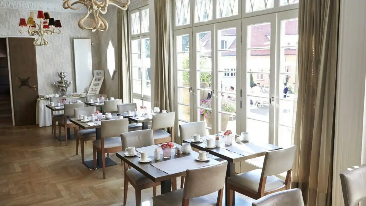 Ein heller Frühstücksraum mit gedeckten Tischen, Blick auf sonnige Terrasse.