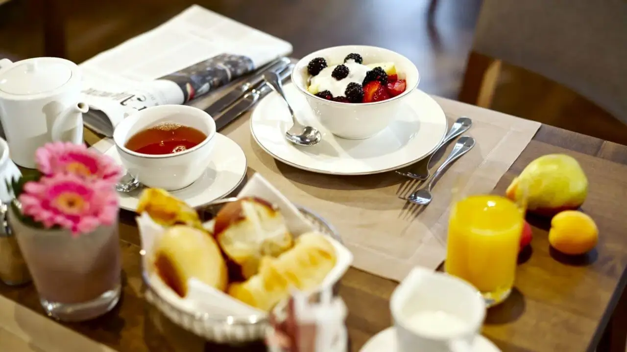 Ein Frühstückstisch mit einer Schale Joghurt und Beeren, einer Tasse Tee, einem Glas Orangensaft, frischem Obst, einem Brötchenkorb, einer Blumenvase und einer Zeitung.