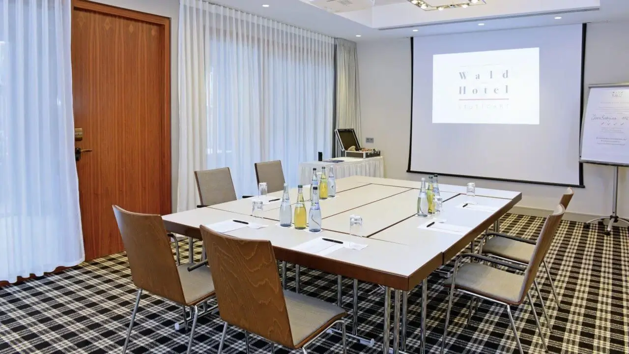 Ein moderner Konferenzraum im Waldhotel Stuttgart mit einem rechteckigen Tisch, Stühlen, Wasserflaschen, Schreibutensilien und einem Projektorbildschirm.