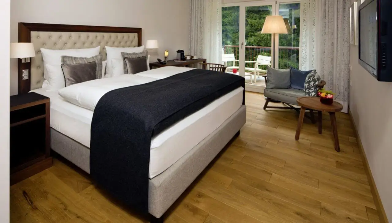 Gemütliches Hotelzimmer mit großem Doppelbett, Holzboden, Schreibtisch einem Sessel mit Beistelltisch und Zugang zu einem Balkon mit Blick ins Grüne.